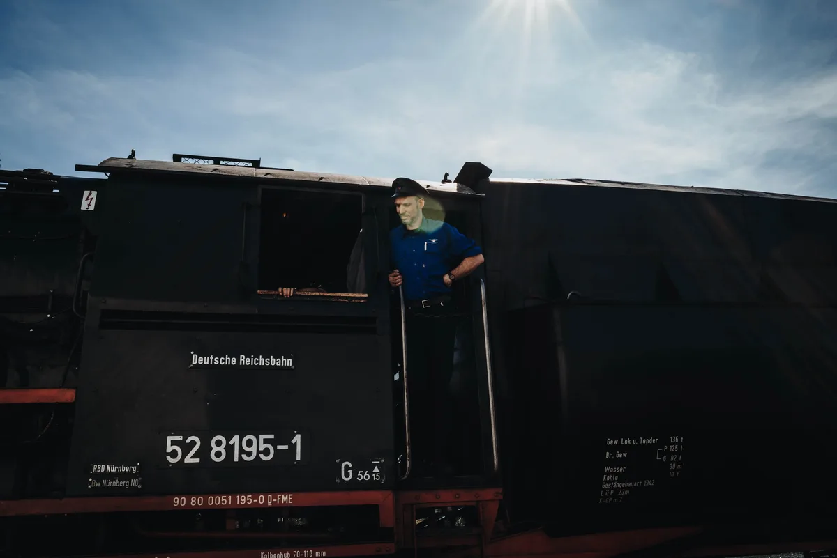 Lokführer in blauer Uniform steht im Gegenlicht in der Tür des Führerhauses, daneben das Schild „Deutsche Reichsbahn“