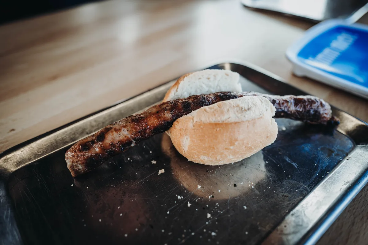 Gegrillte Bratwurst liegt auf einem aufgeschnittenen Brötchen auf einem Edelstahltablett, daneben eine blaue Schale