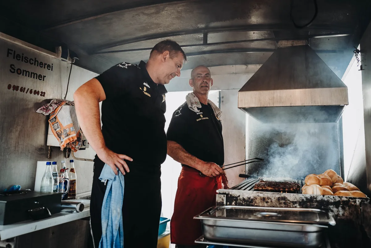 Zwei Männer in schwarzen Shirts stehen am Grillwagen, einer wendet mit der Zange Würste, daneben Brötchen und Dampf