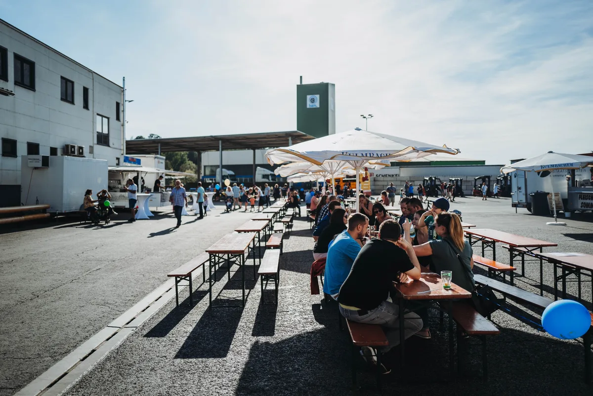 Gäste sitzen an Biertischgarnituren unter weißen Sonnenschirmen, links Gebäude und Imbissstände, rechts ein blauer Luftballon