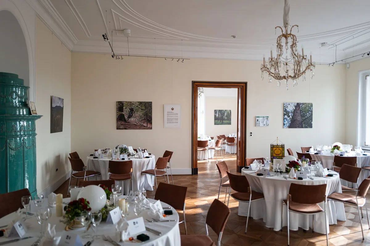 Festlich gedeckte runde Tische in einem hohen Saal mit Kronleuchter, grünem Kachelofen und Waldfotografien an der Wand
