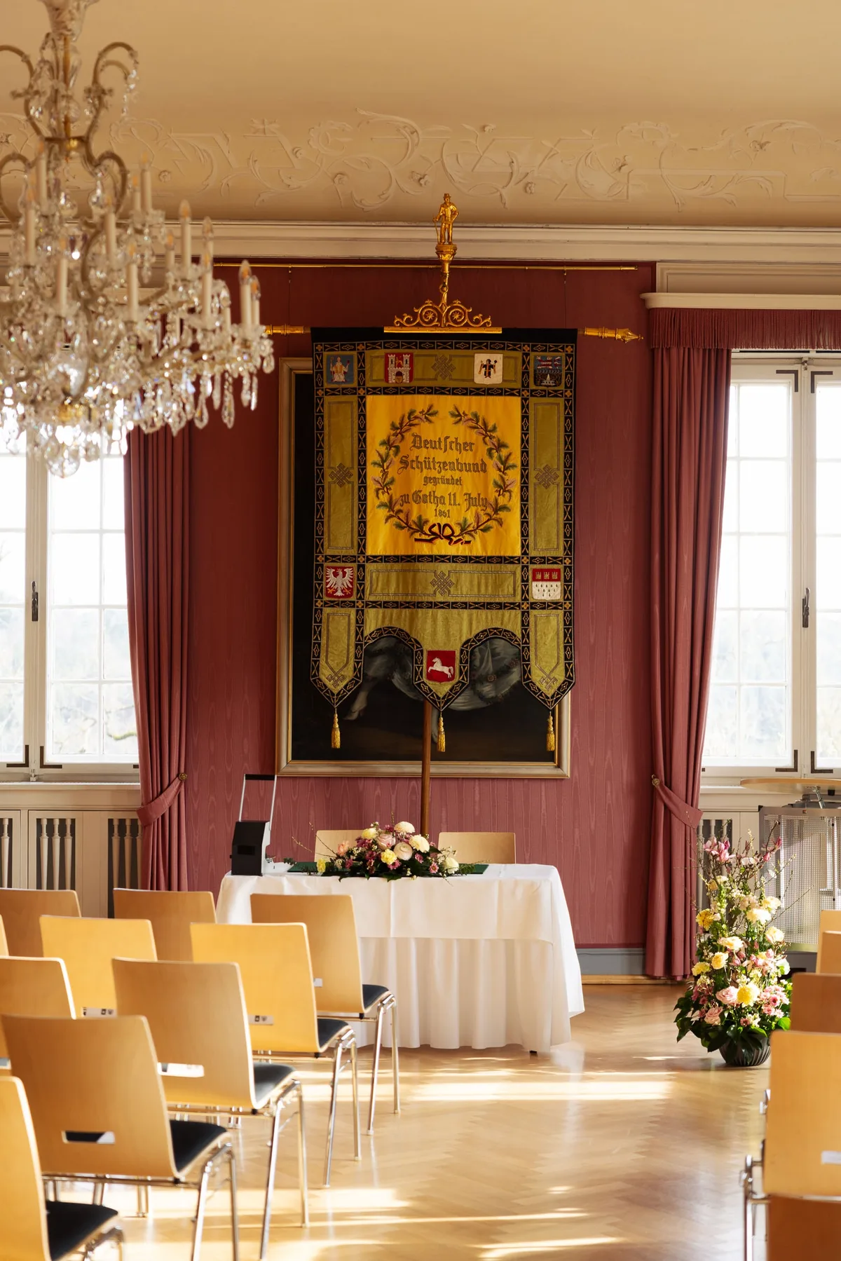 Hochformat: Bestickte Fahne über einem Tisch mit Blumengesteck, davor leere Stuhlreihen im Saal mit roten Wänden