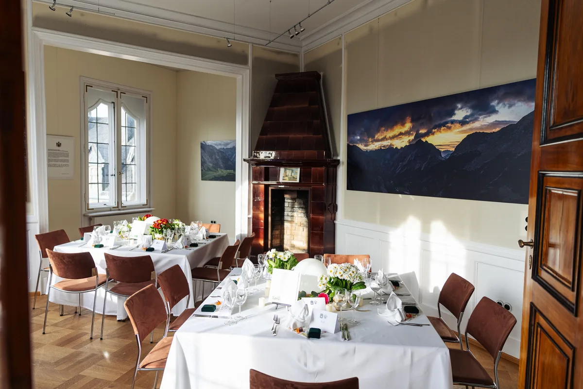 Kleiner Raum mit langer eingedeckter Tafel, braunem Kachelofen und einem Bergpanorama im Abendrot an der Wand