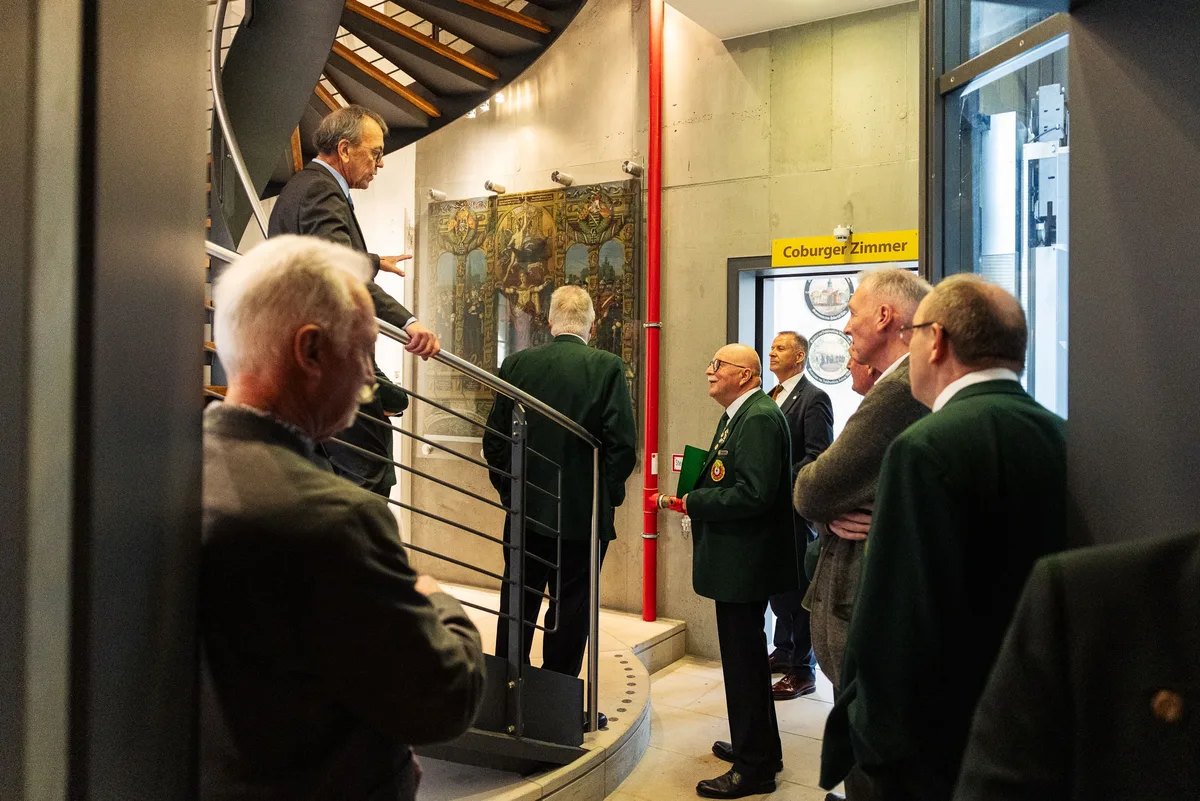 Gäste in grünen Jacken und Anzügen steigen an einer Wendeltreppe vor einem Gemälde an der Betonwand zusammen