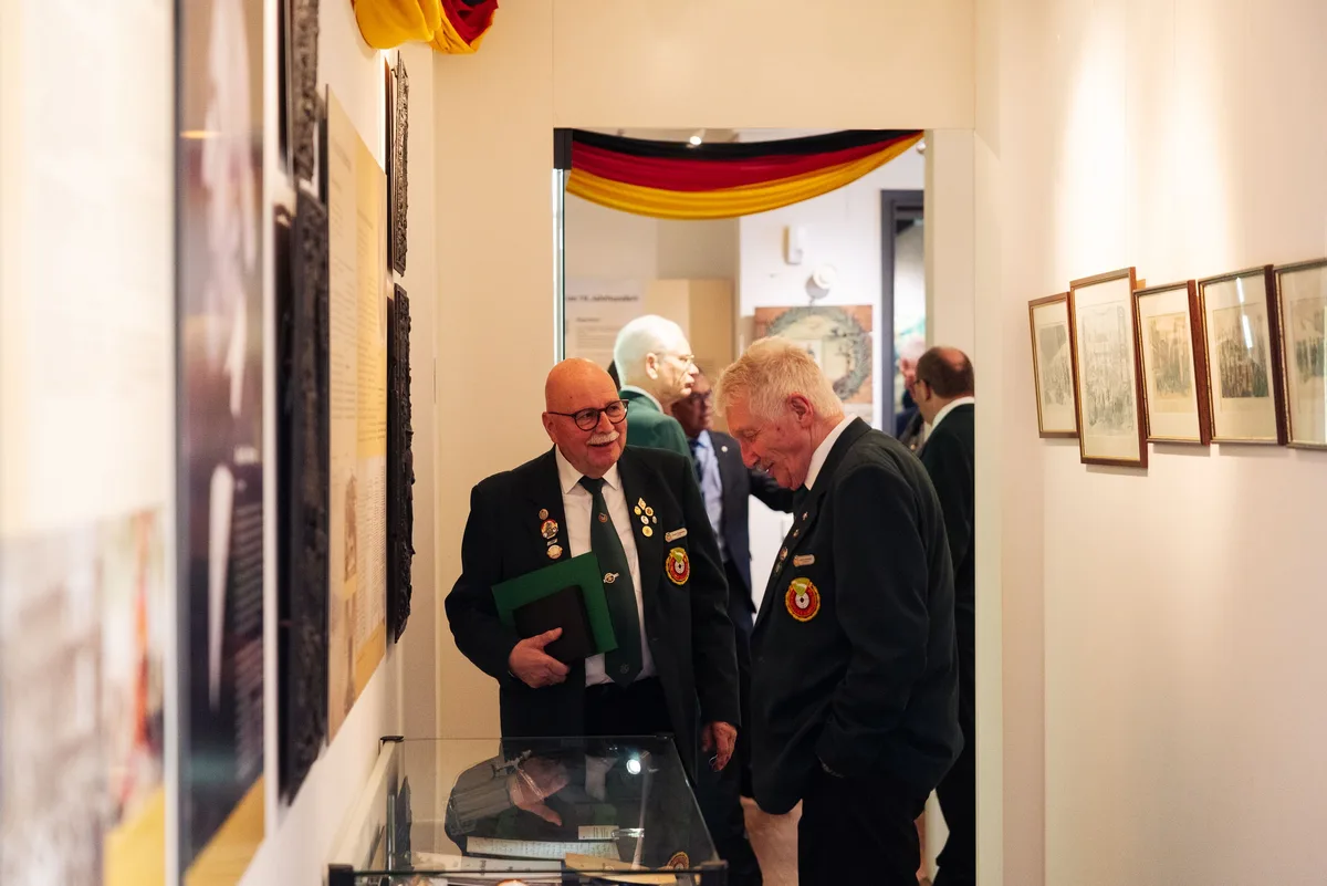 Zwei Männer in dunklen Jacken mit Abzeichen im Gespräch in einem Ausstellungsgang, hinten ein Fahnenband in Schwarz-Rot-Gold