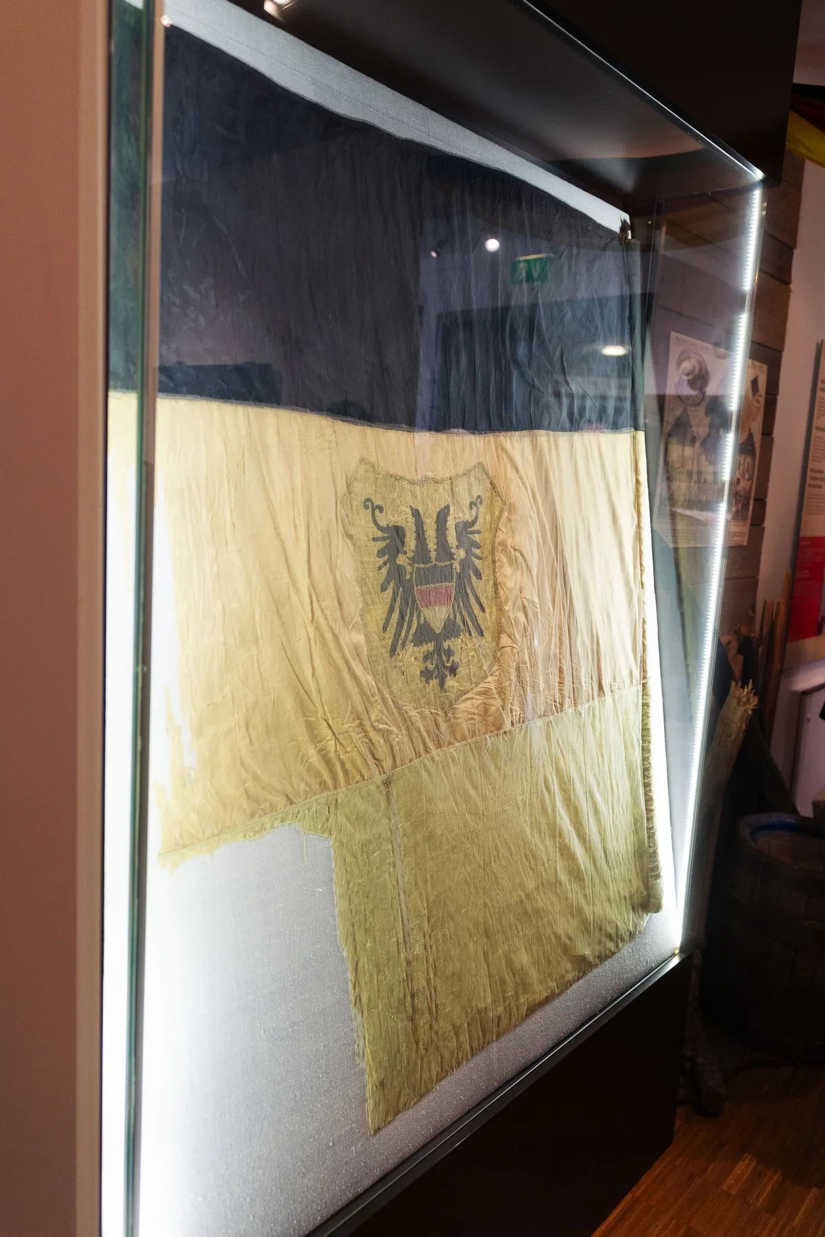 Hochformat: Alte vergilbte Fahne mit Doppeladler in einer beleuchteten Glasvitrine