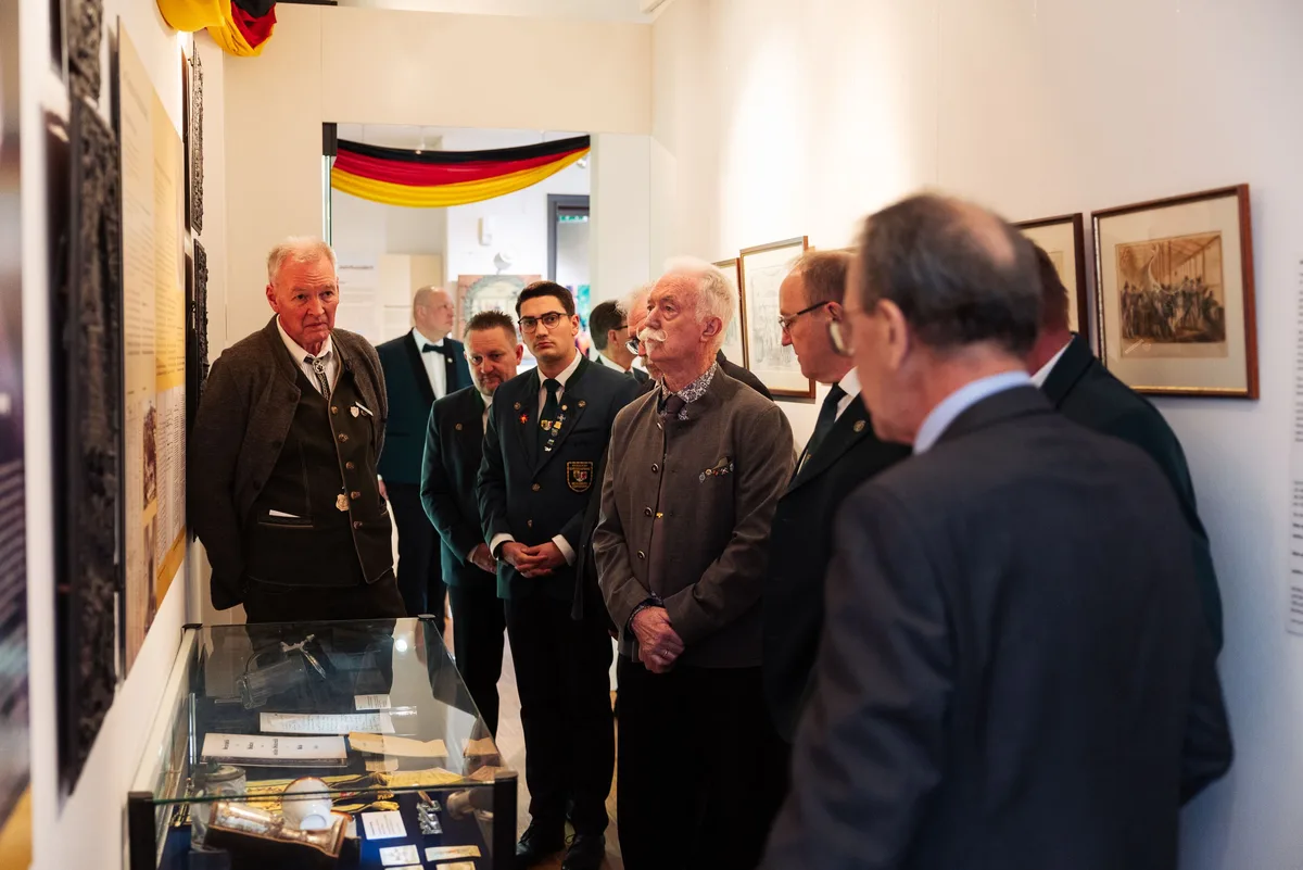 Mehrere Gäste, teils in Schützenjacken, stehen im Ausstellungsgang vor einer Vitrine und gerahmten Grafiken