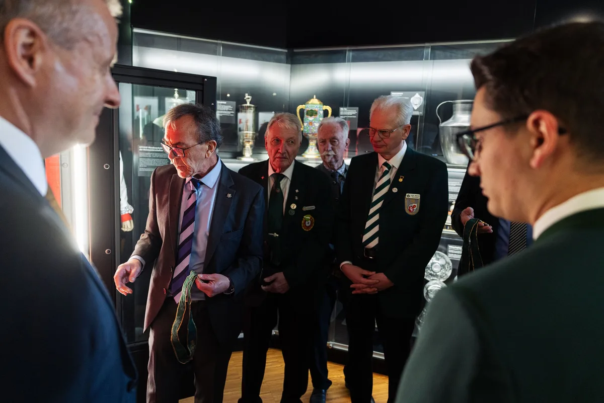 Mann im Anzug hält ein grün gemustertes Band und erklärt einer Gruppe vor einer Vitrine mit Pokalen etwas