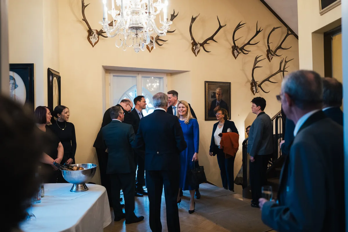 Gäste im Treppenhaus mit Hirschgeweihen an der Wand und Kronleuchter, eine Frau im blauen Kleid mittendrin
