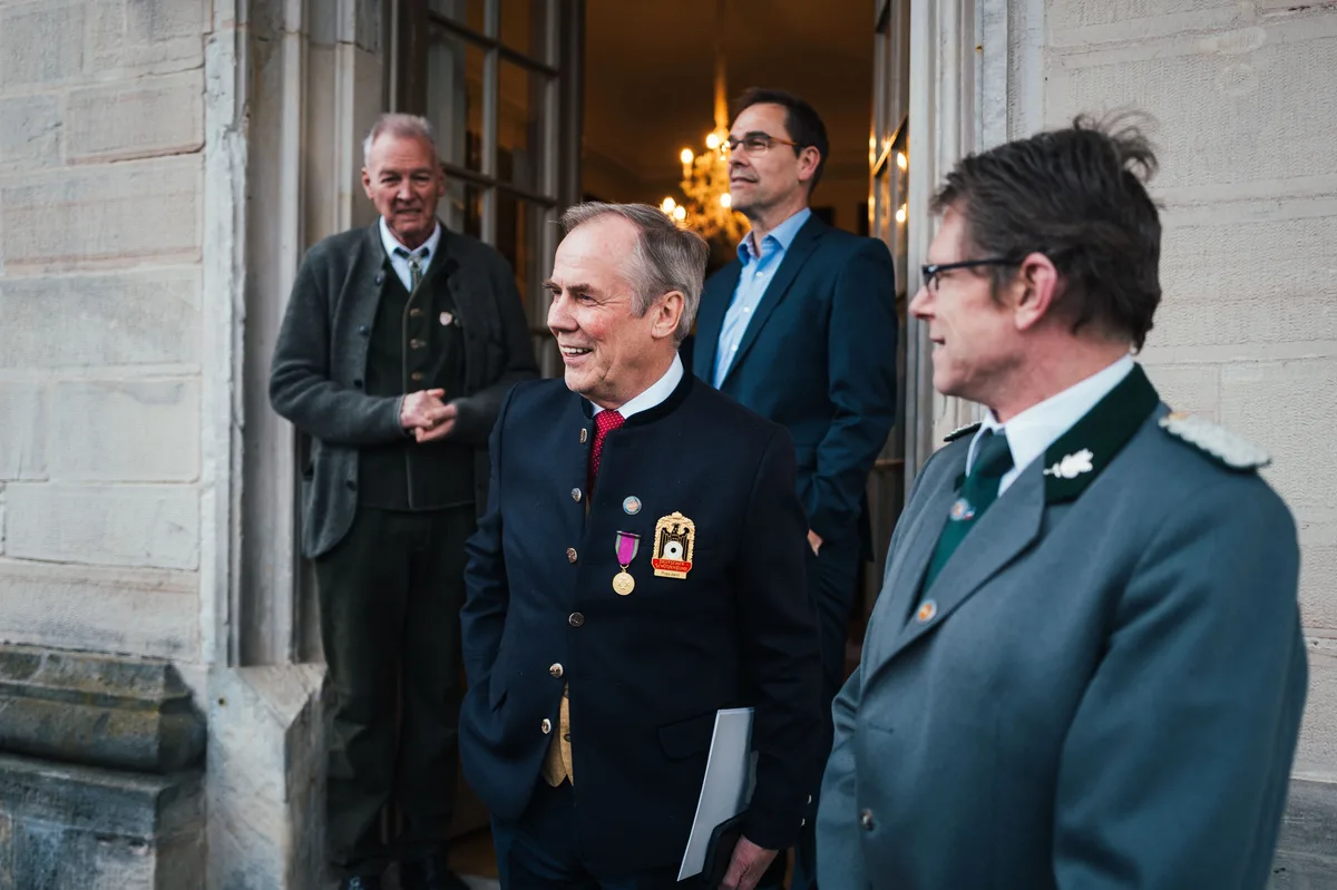 Mann mit Orden und Mappe lacht, daneben ein Mann in grauer Uniform mit Schulterstücken, hinten ein weiterer am Portal