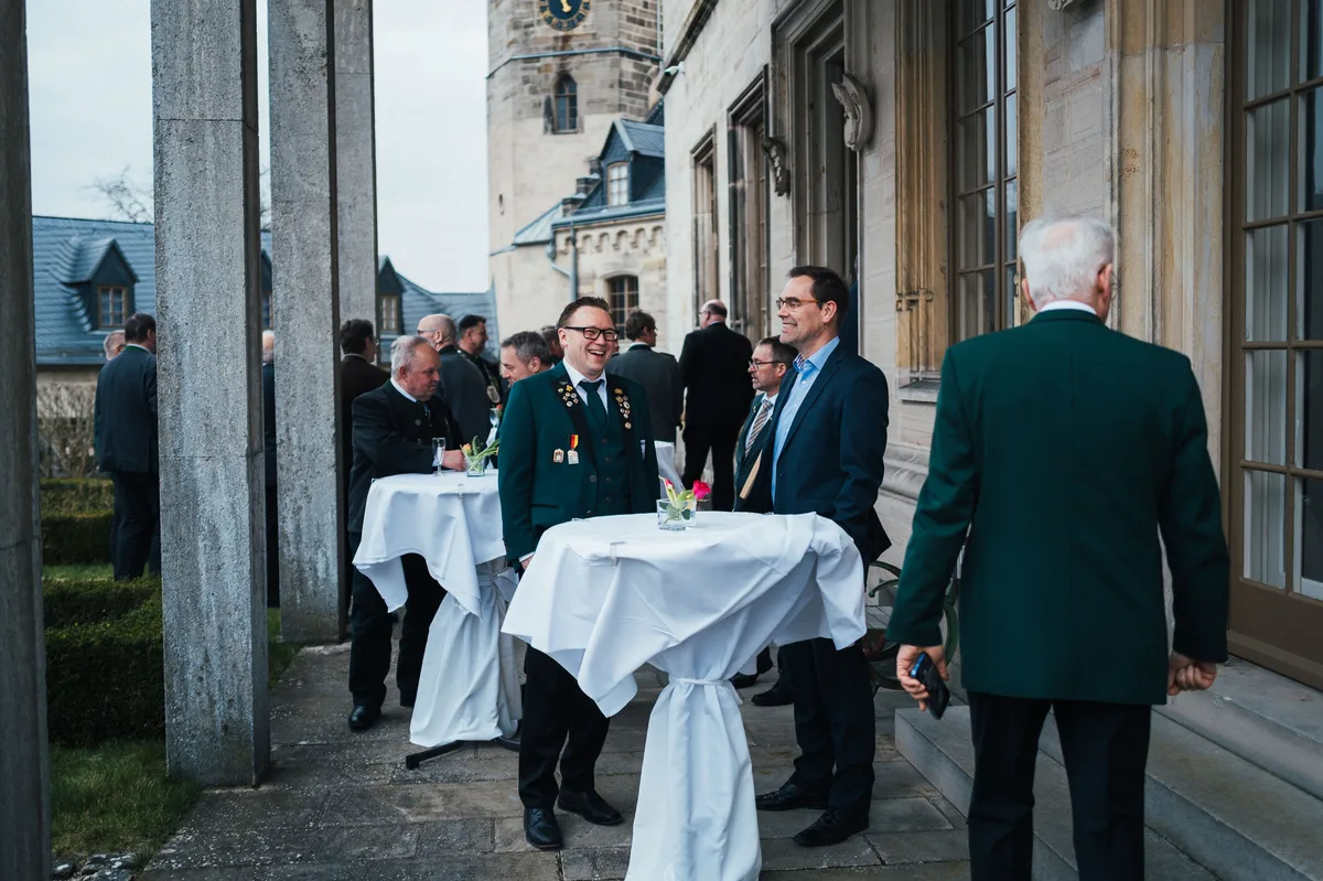 Stehtische mit weißen Hussen auf der Terrasse, ein Mann mit Brille in grüner Jacke lacht, dahinter der Uhrturm