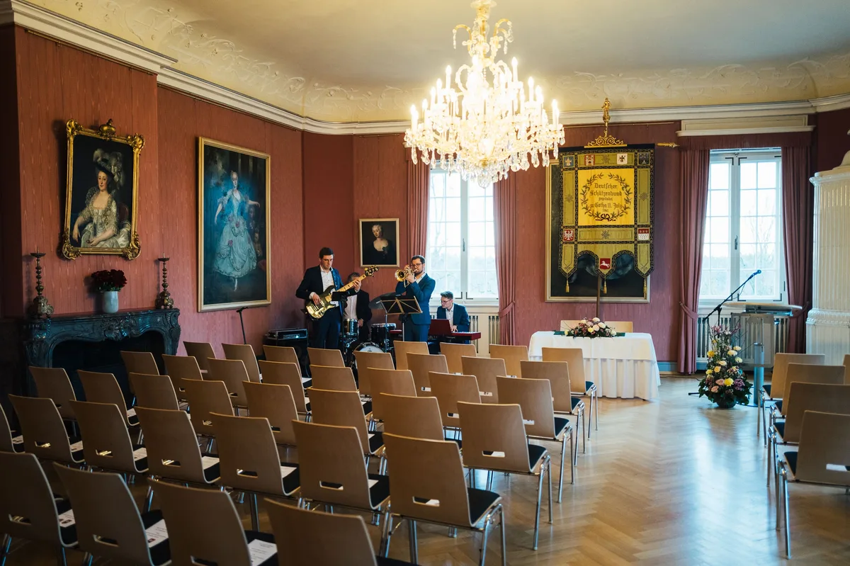 Dreiköpfige Band mit Bass, Trompete und Keyboard vor leeren Stuhlreihen, links gerahmte Porträts, rechts die Fahne