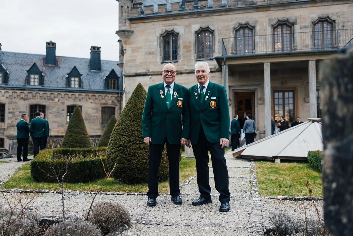 Zwei lächelnde Männer in grünen Schützenjacken im Schlossgarten, hinter ihnen Buchsbaumkegel und Schlossfassade