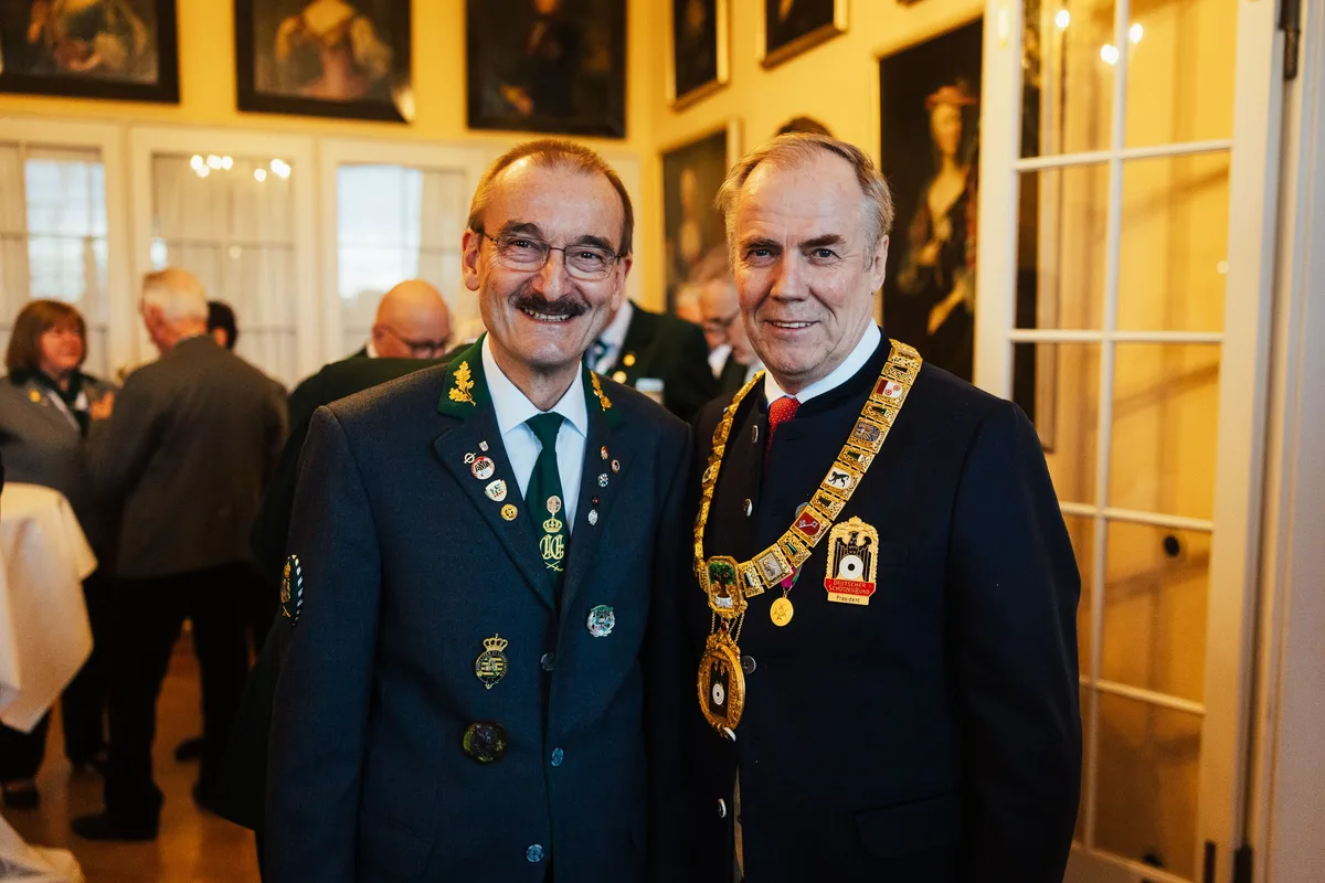 Mann in grüner Jacke mit vielen Abzeichen neben einem Mann mit goldener Amtskette, im Hintergrund Gäste im Saal