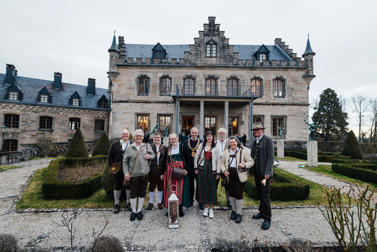 Gruppe in bayerischer Tracht mit Laterne posiert im Garten, in der Mitte ein Mann mit Amtskette, dahinter das Schloss
