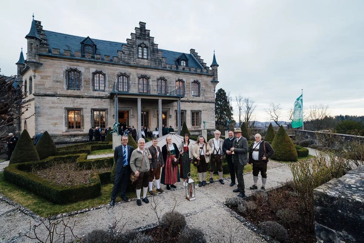 Gruppe in Tracht mit Laterne im Garten zwischen Buchsbaumhecken, rechts weht eine grüne Fahne