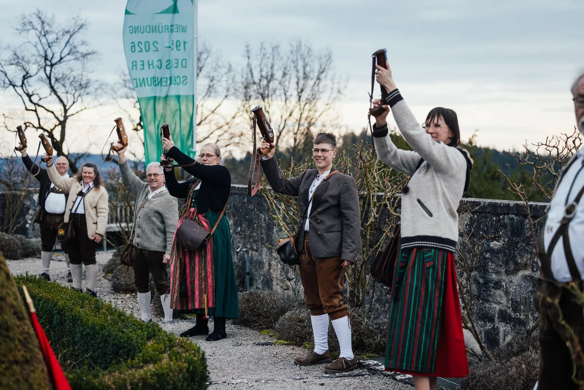 Menschen in Tracht heben Vorderladerpistolen in die Luft, links ein Schützenbund-Banner vor kahlen Bäumen
