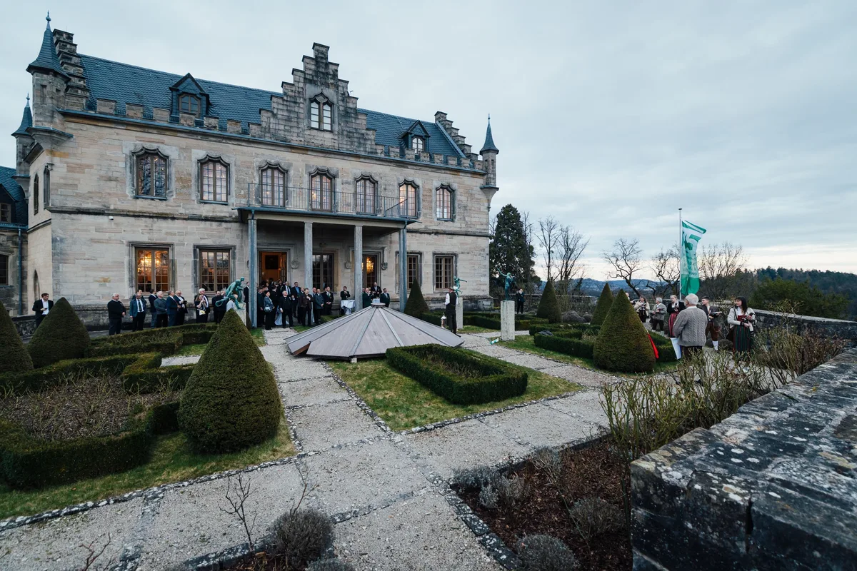 Weitwinkel vom Garten aus: Gäste vor dem Schlossportal, Buchsbaumhecken, rechts die grüne Fahne und Abendhimmel