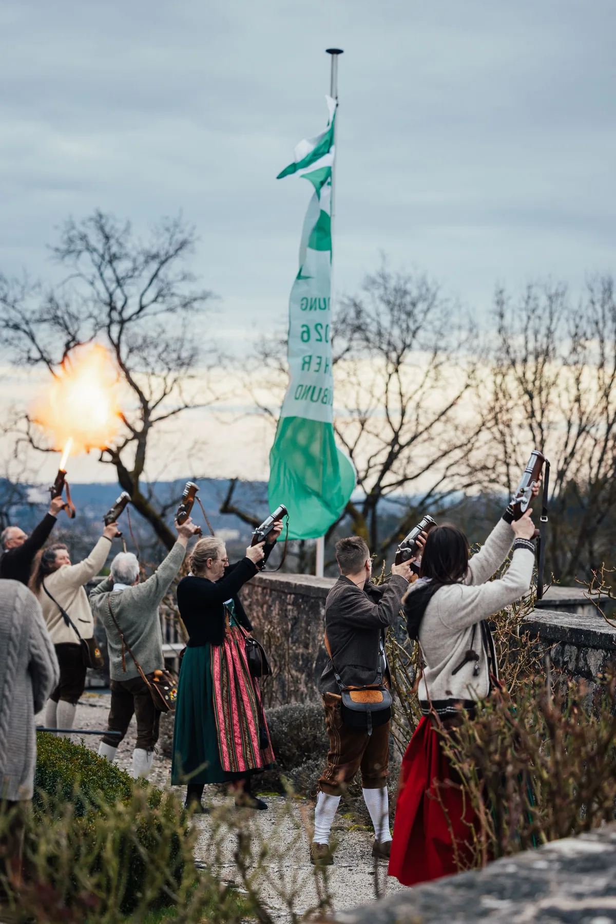 Hochformat: Tracht tragende Schützen feuern Salut, links schießt eine Flamme aus einer Pistole, in der Mitte eine Fahne