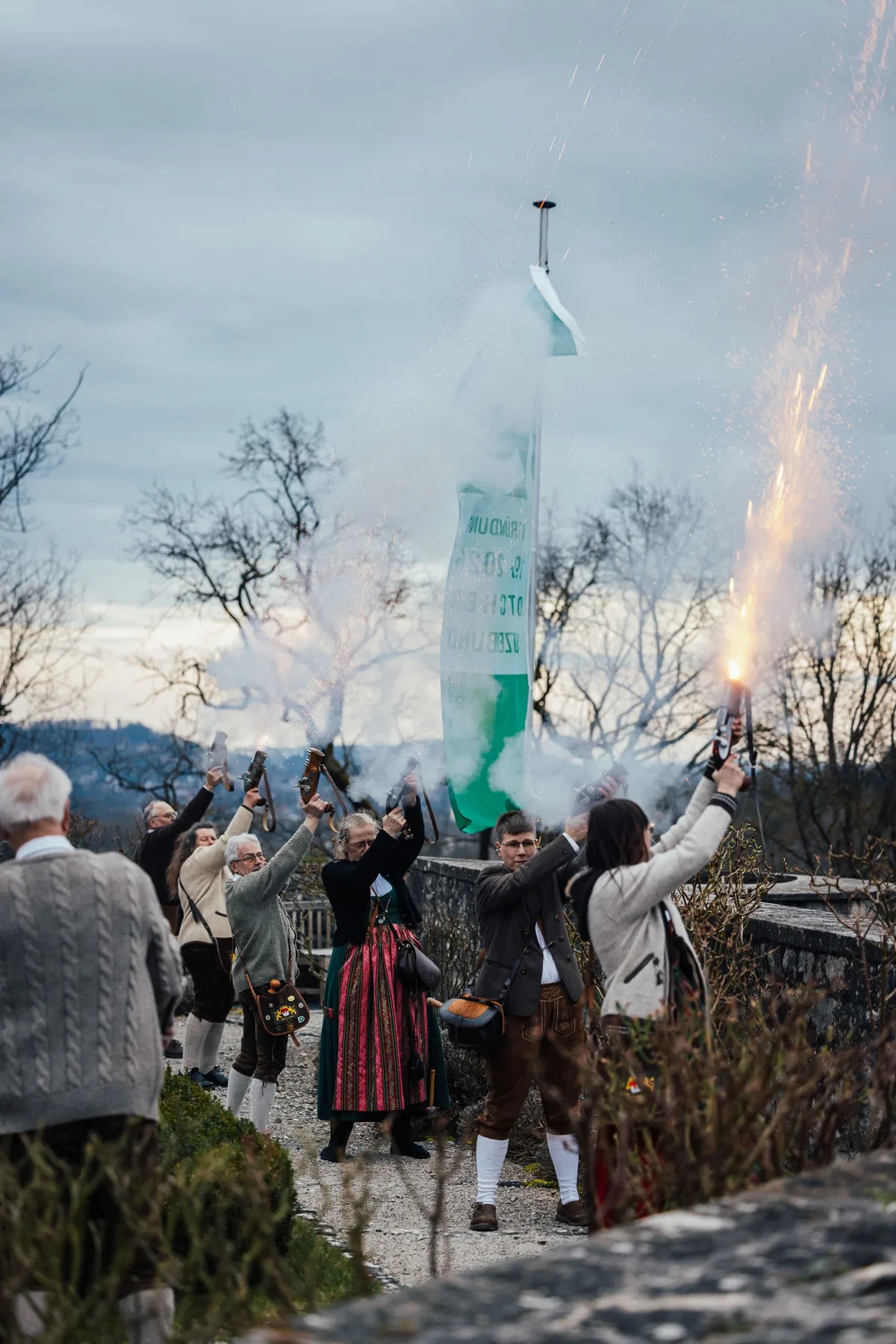 Hochformat: Salutschützen im Garten, rechts sprühen Funken aus einer Pistole, Rauch zieht vor der Fahne und kahlen Bäumen