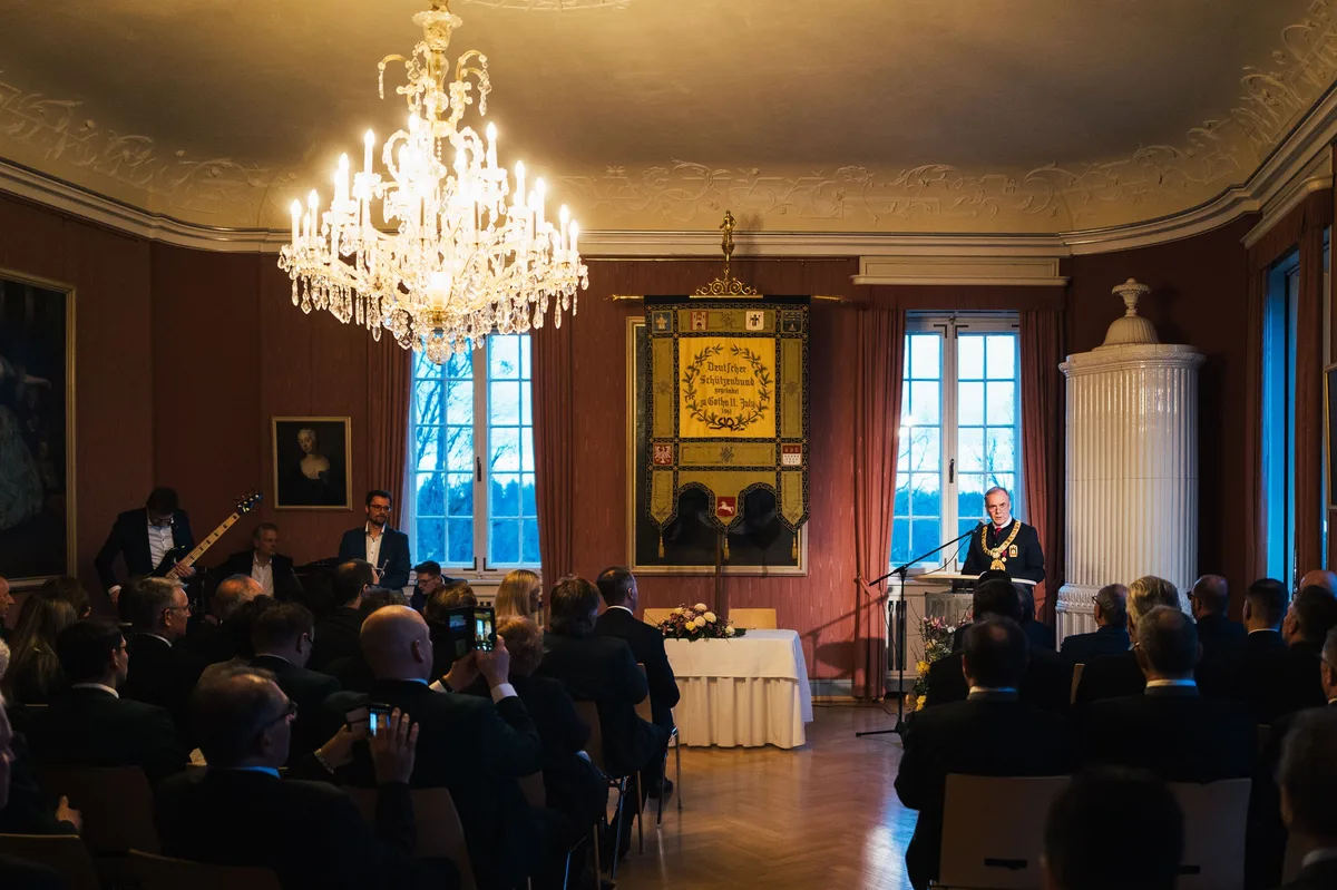 Festsaal mit Kronleuchter, Publikum im Vordergrund, am Rednerpult ein Mann mit Amtskette, links spielt die Band