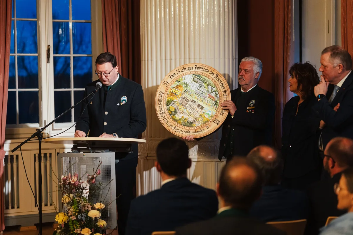 Redner am Pult, neben ihm hält ein Mann einen runden Holzteller mit Schlossmotiv, dahinter Gäste im Halbdunkel