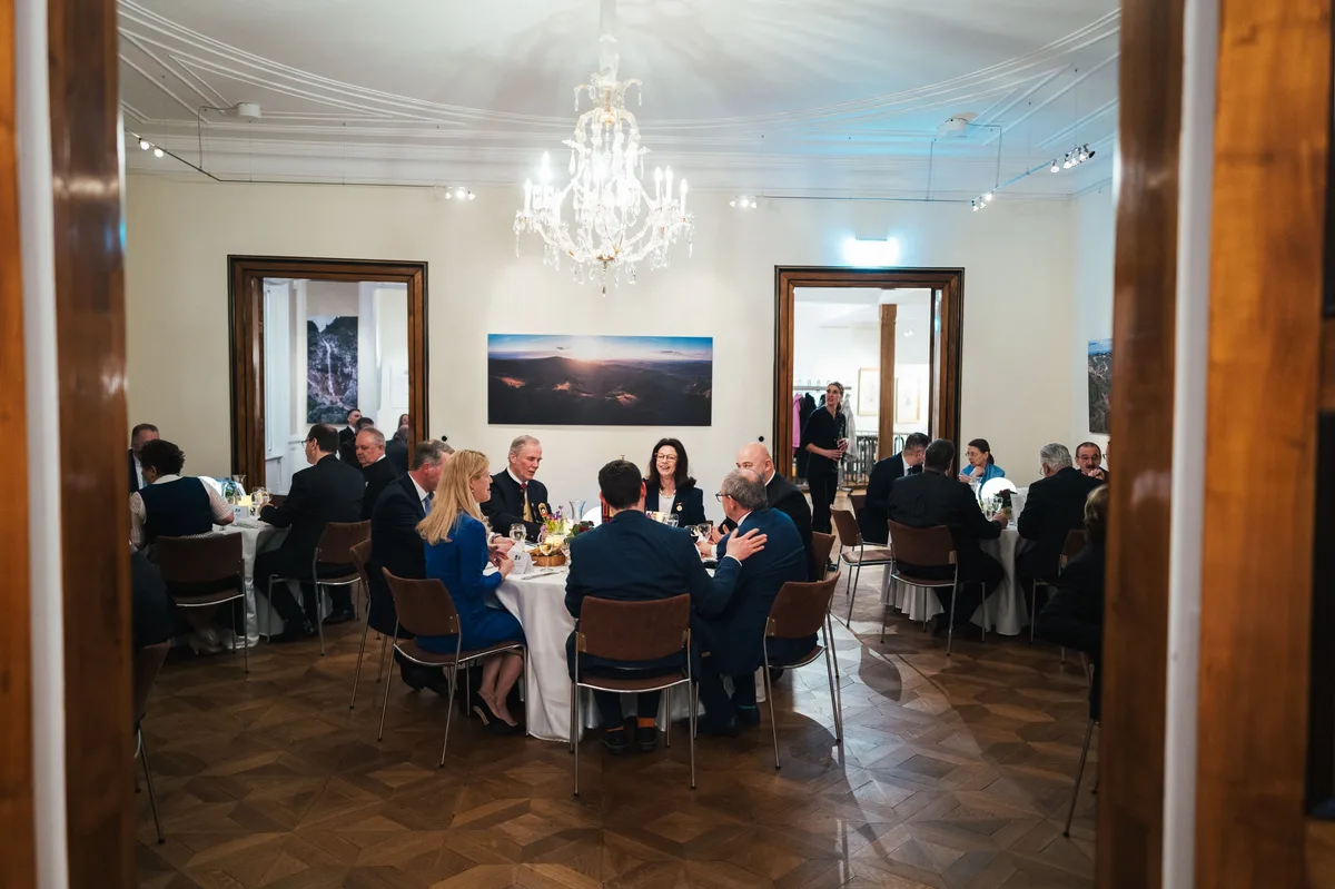 Blick durch eine Tür auf Gäste an einem runden Tisch unter dem Kronleuchter, an der Wand ein Panoramabild