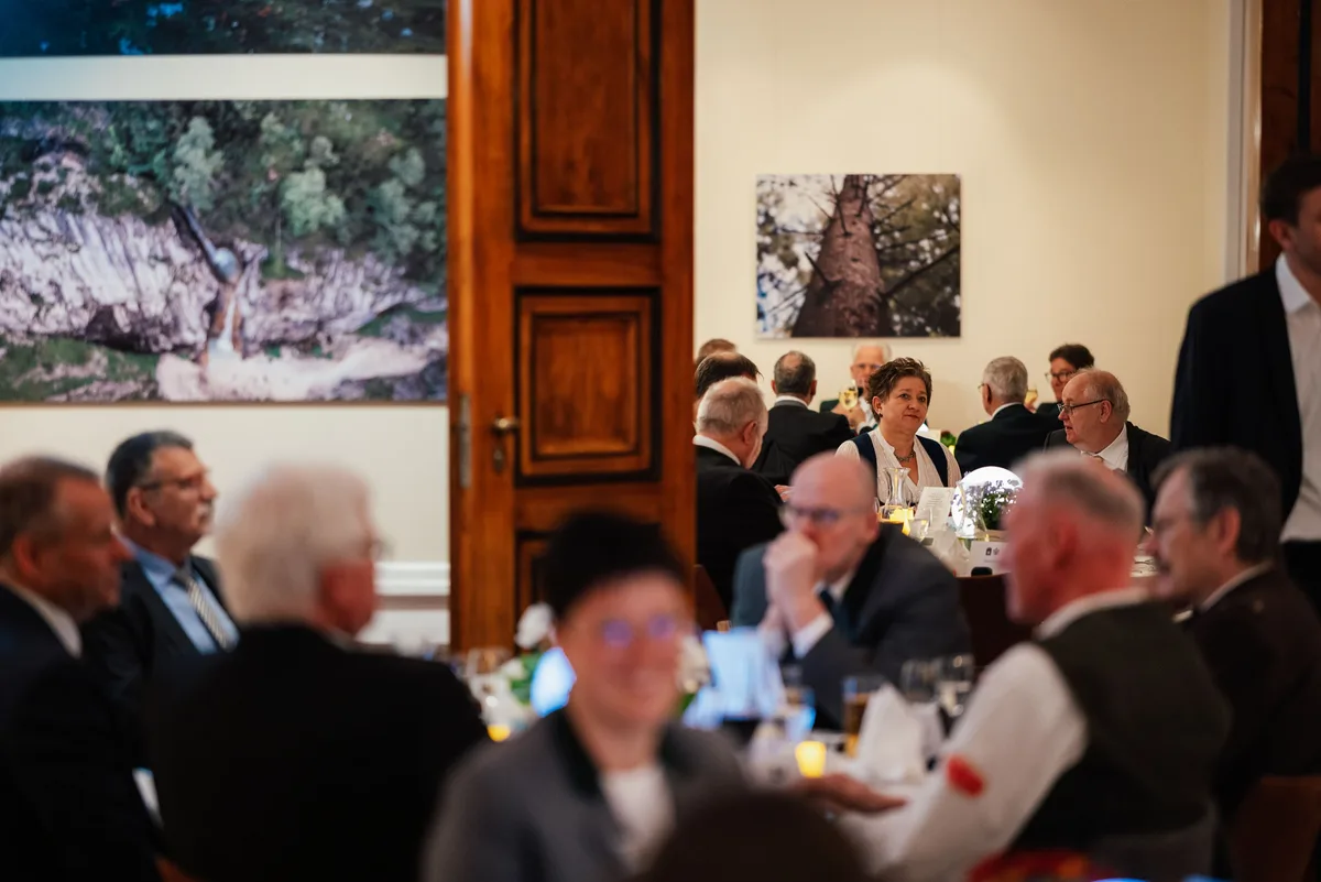 Unscharf im Vordergrund Gäste am Tisch, im Hintergrund eine lächelnde Frau am Tisch und ein Baumfoto an der Wand