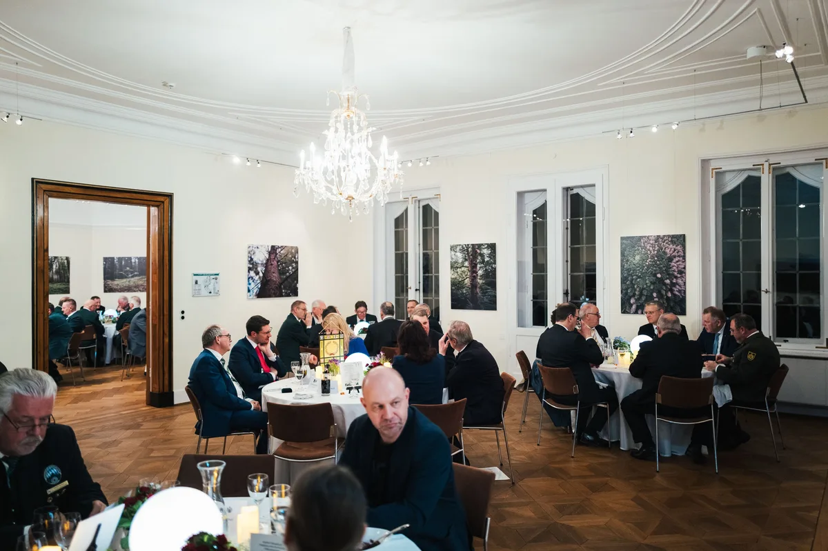 Saal mit mehreren besetzten Tischen unter dem Kronleuchter, an der Wand Waldfotos, im Vordergrund ein Mann mit Glatze