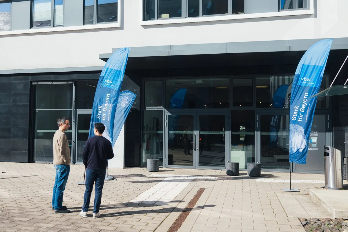 Zwei Männer im Gespräch vor einem Gebäudeeingang, links und rechts blaue Beachflags mit der Aufschrift „Stark für Bayern“