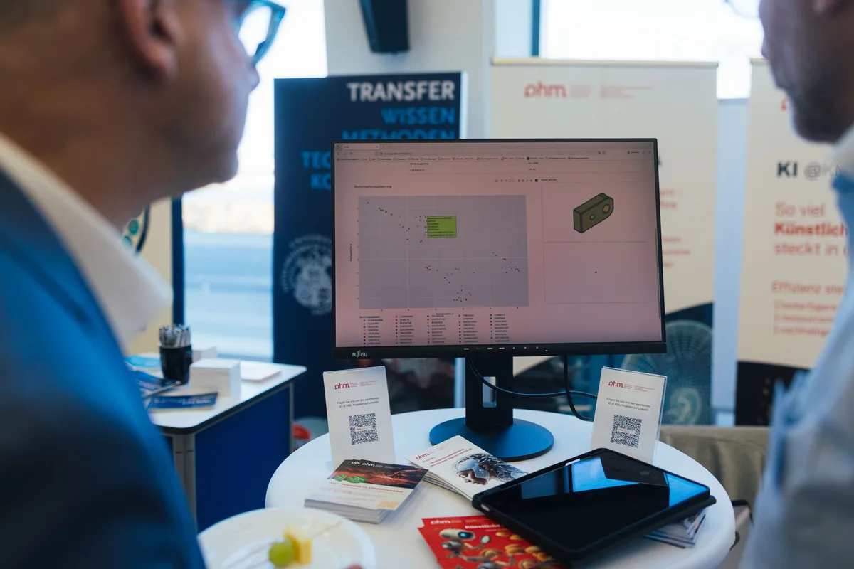 Ausstellungstisch mit Monitor, der ein Streudiagramm und ein 3D-Bauteil zeigt, davor zwei Gesprächspartner im Anschnitt