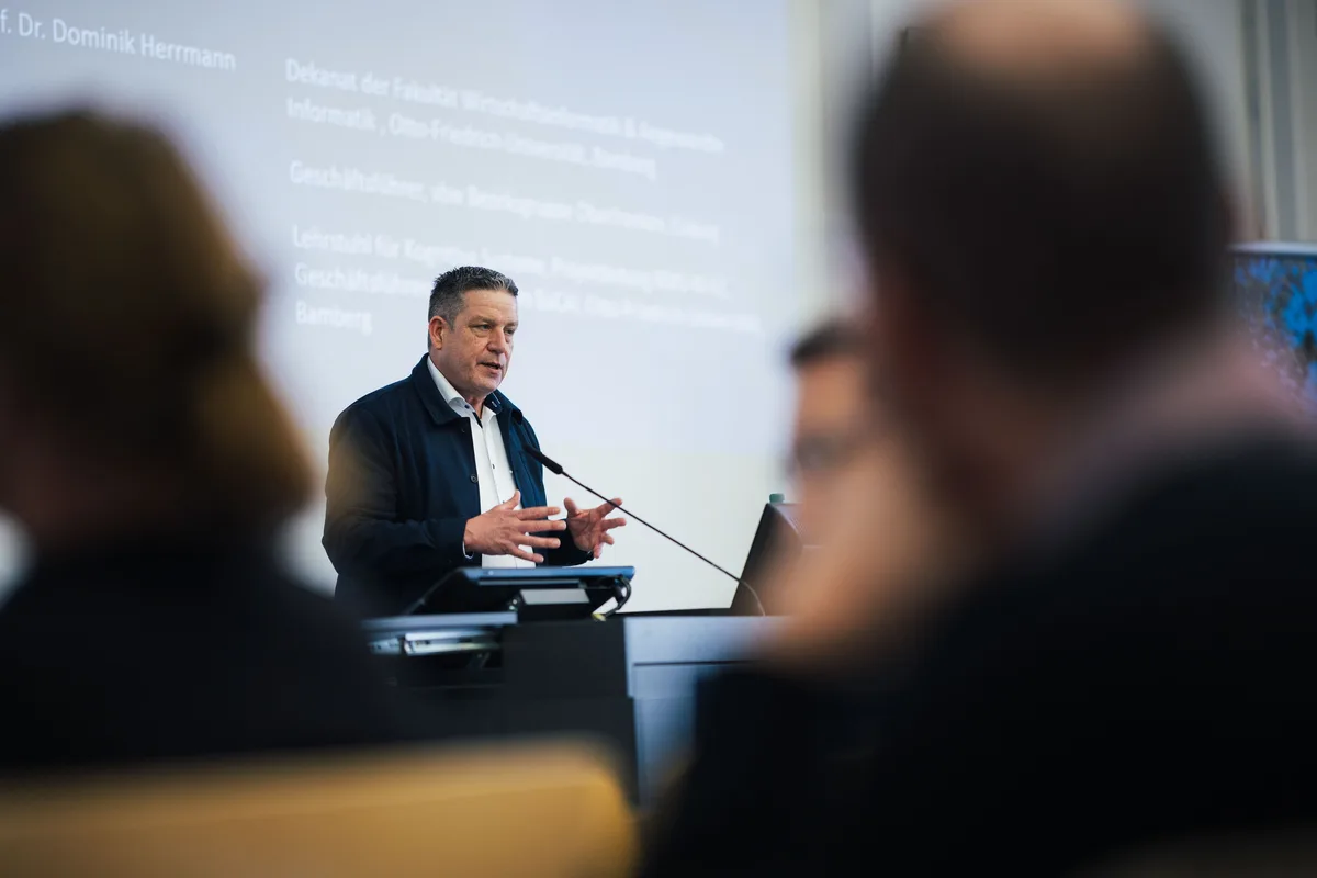 Mann im Sakko spricht mit ausgebreiteten Händen am Pult, im Vordergrund große unscharfe Köpfe, dahinter die Projektion