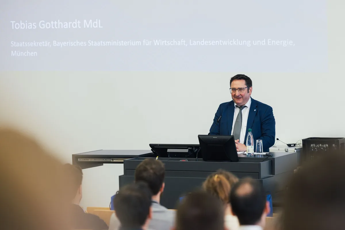 Mann im blauen Anzug mit Brille spricht lächelnd am Pult unter der Projektion, davor unscharfe Köpfe des Publikums