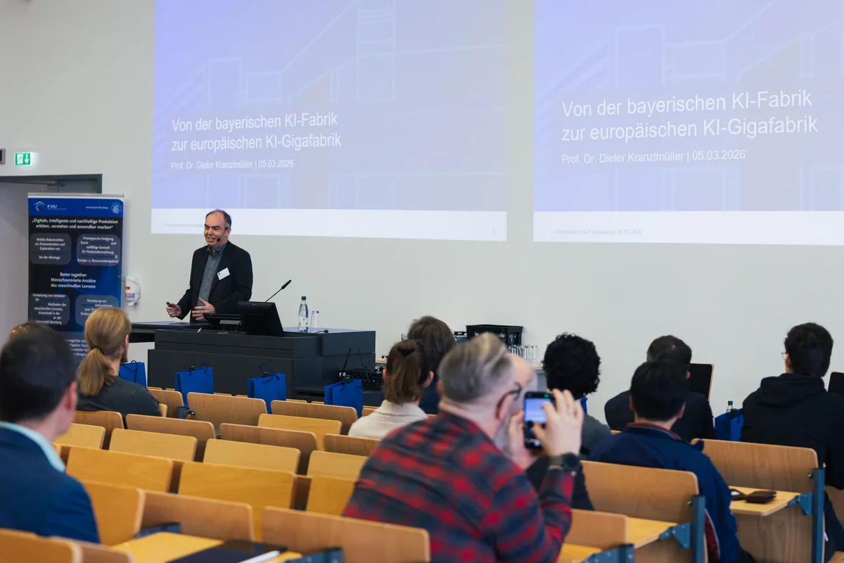 Redner am Pult vor zwei Projektionen zur europäischen KI-Gigafabrik, davor Zuhörer, einer fotografiert mit dem Handy