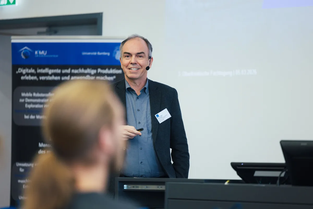 Redner mit Headset und Namensschild lächelt, in der Hand einen Presenter, davor unscharf ein Zuhörer mit Pferdeschwanz