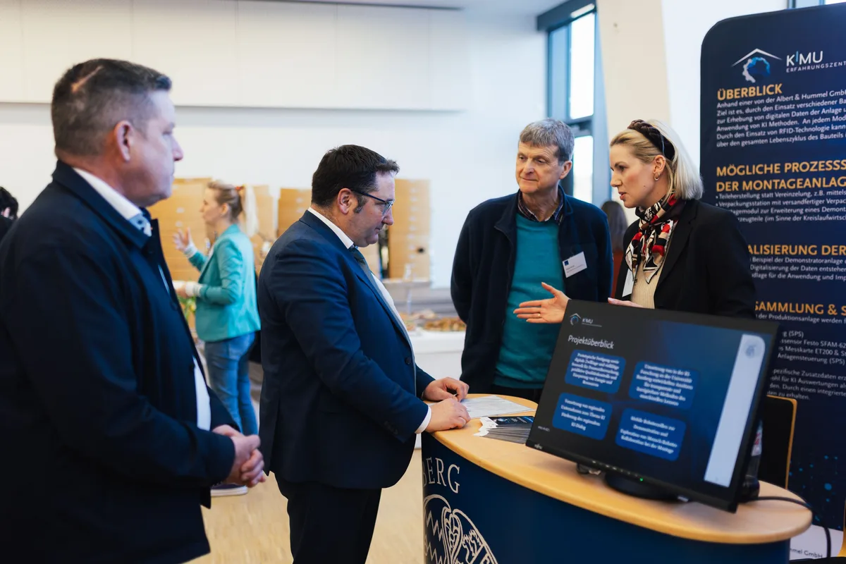 Vier Männer und eine Frau im Gespräch am Messestand, ein Mann im Anzug beugt sich über den Tisch, rechts ein Monitor