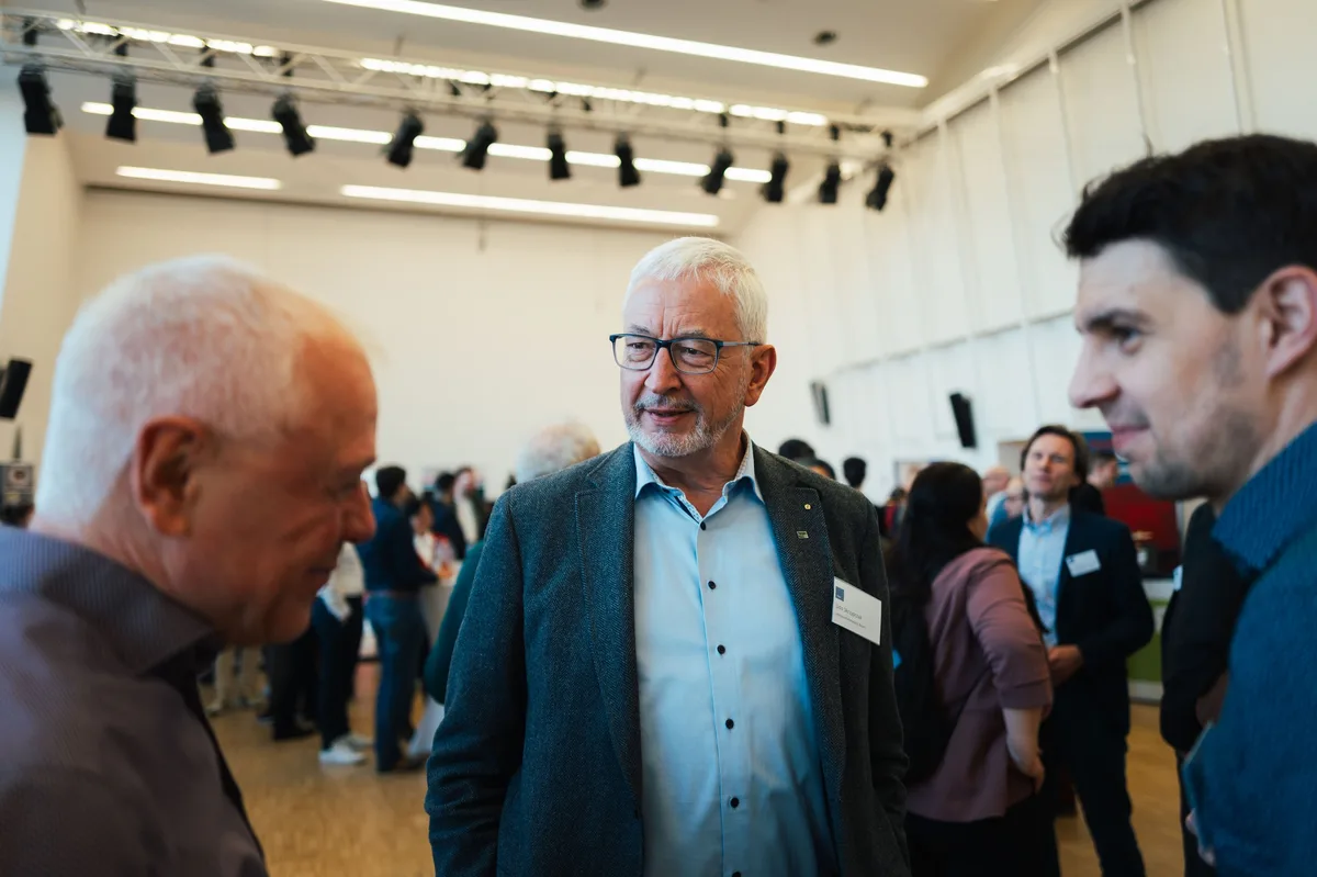 Älterer Mann mit Brille und grauem Sakko im Gespräch, links und rechts Gesprächspartner im Anschnitt, hinten Gäste