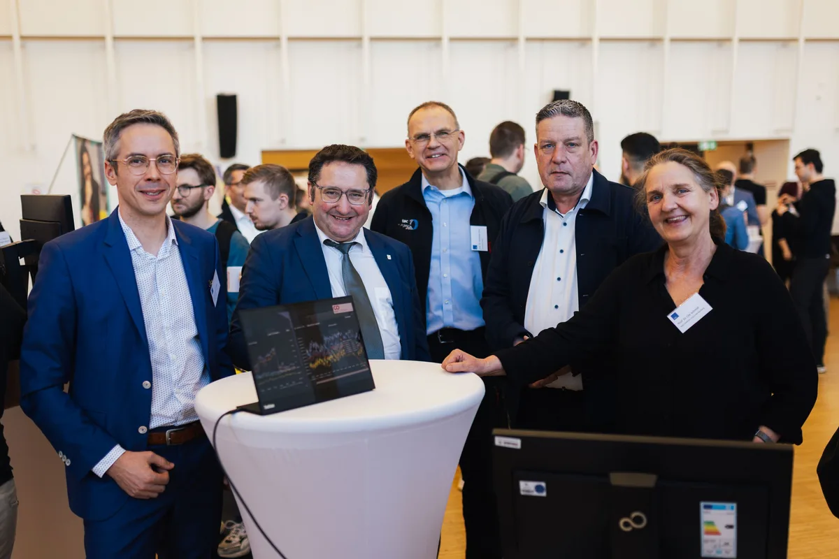 Fünf Personen stehen lächelnd an einem Stehtisch mit Laptop, rechts im Vordergrund ein Monitor, dahinter Gäste im Foyer