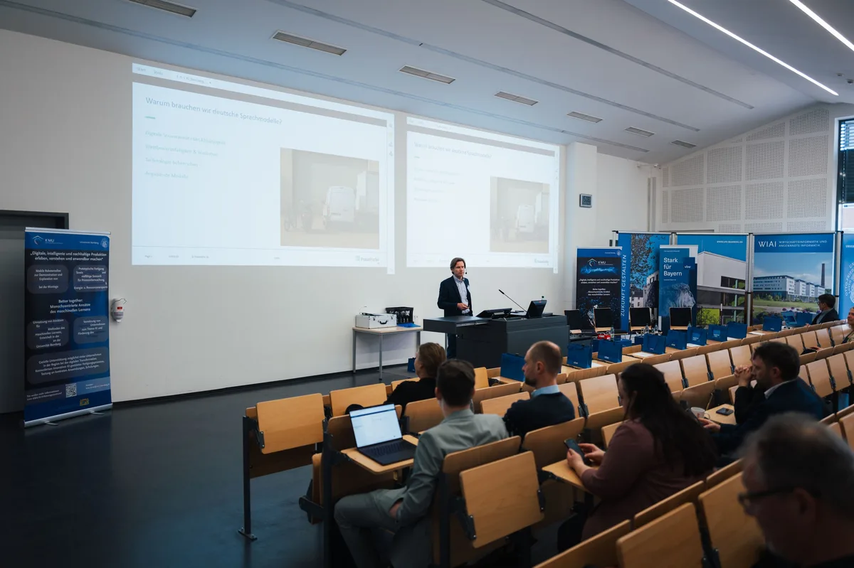 Blick durch den Hörsaal auf den Redner am Pult, zwei Projektionsflächen zeigen Folien, davor Zuhörer mit Laptop und Handy