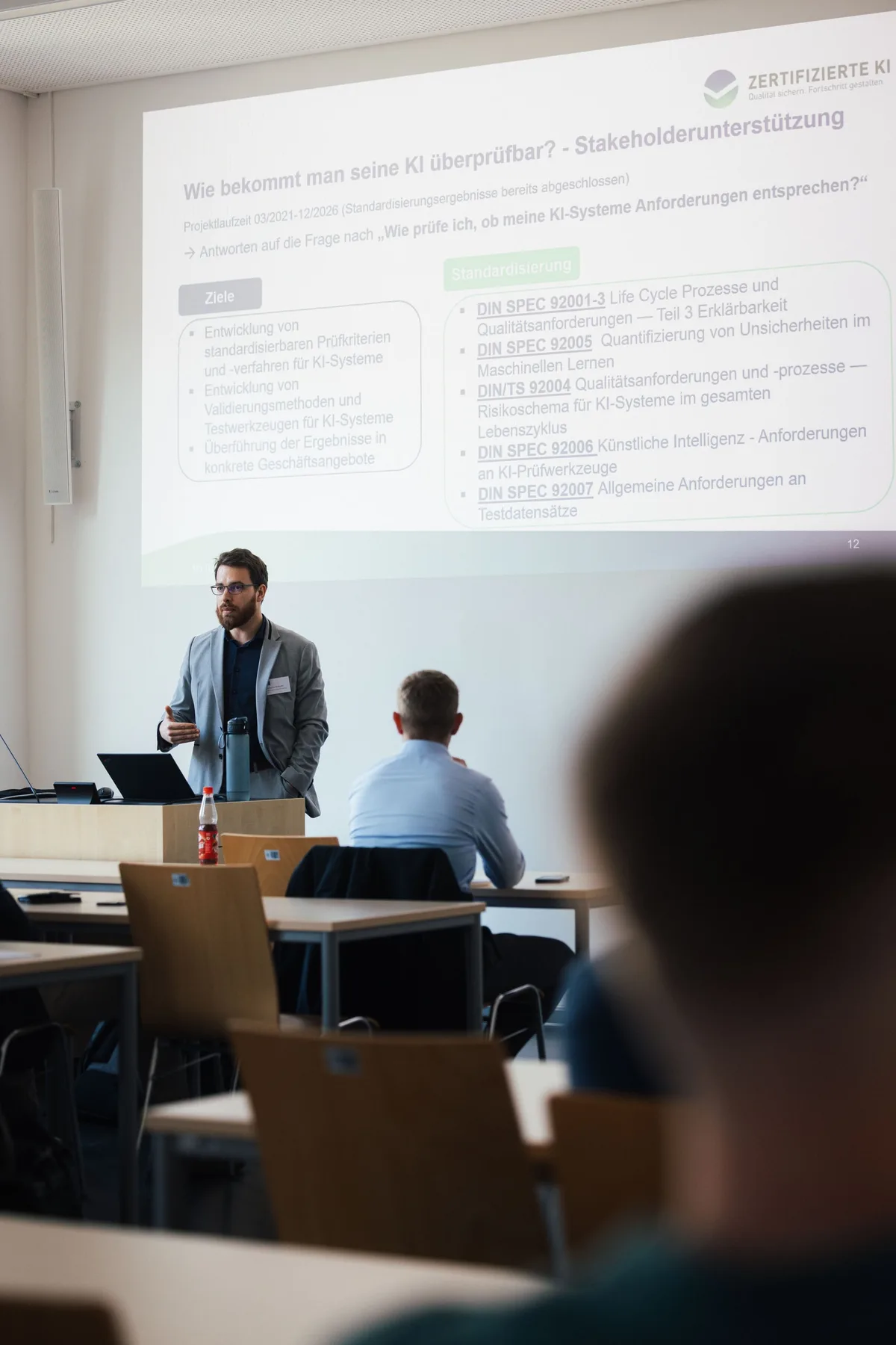 Hochformat: Referent im grauen Sakko erklärt vor einer Folie zur Prüfbarkeit von KI, davor unscharf ein Kopf von hinten