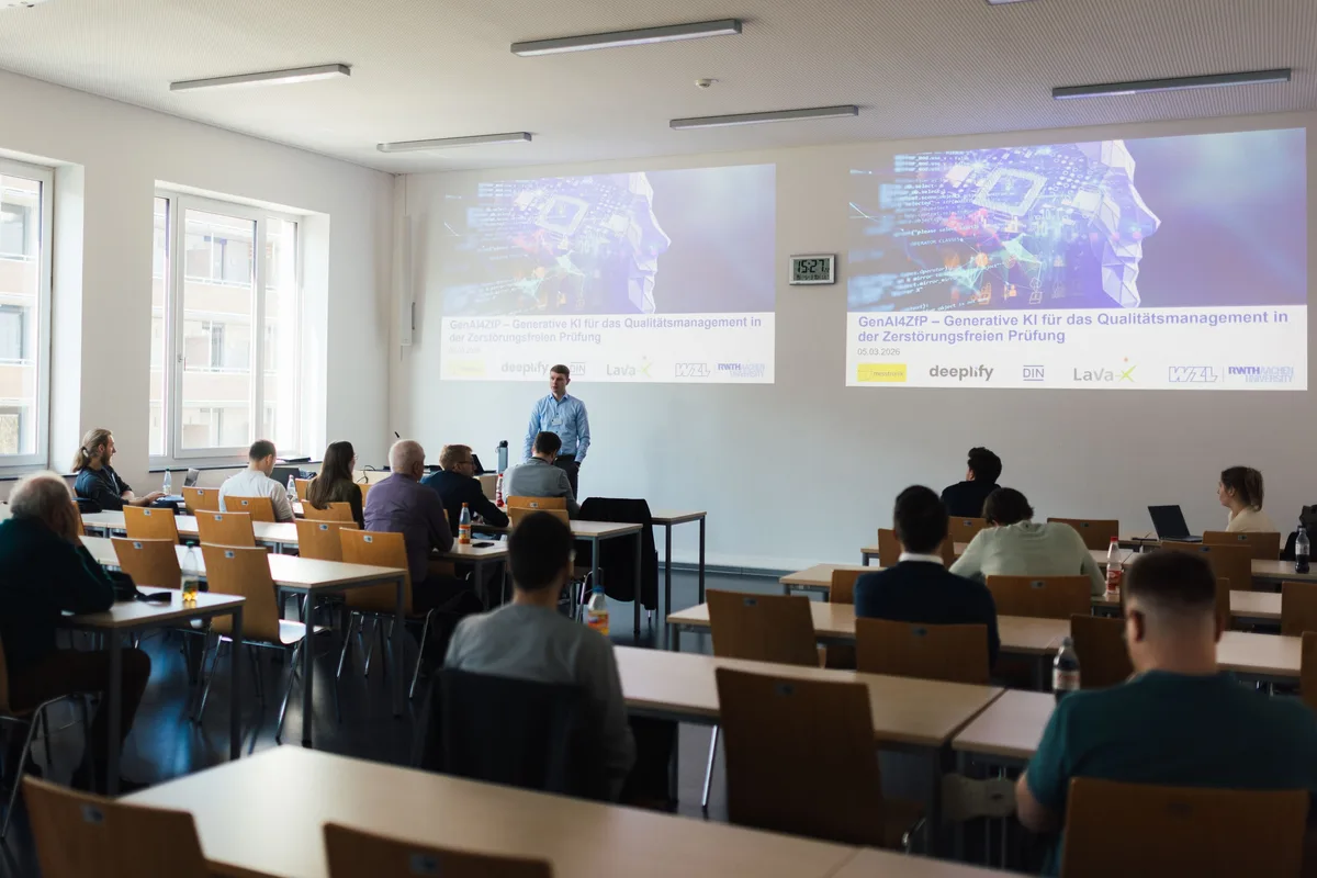 Referent im blauen Hemd steht im Seminarraum vor zwei Projektionen, an den Tischreihen sitzen verteilt einige Teilnehmende