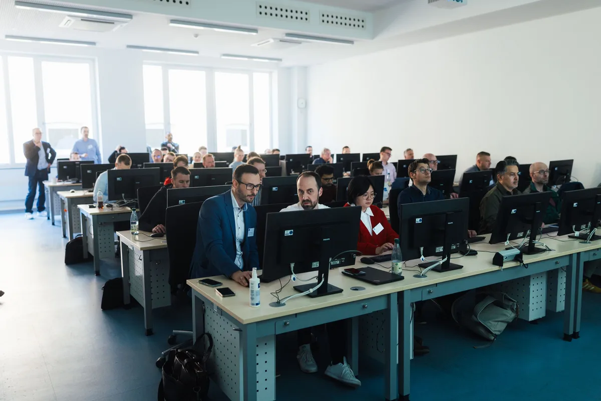 Teilnehmende sitzen in Reihen vor Monitoren im Computerraum, vorn ein Mann im blauen Sakko, hinten stehen zwei Personen