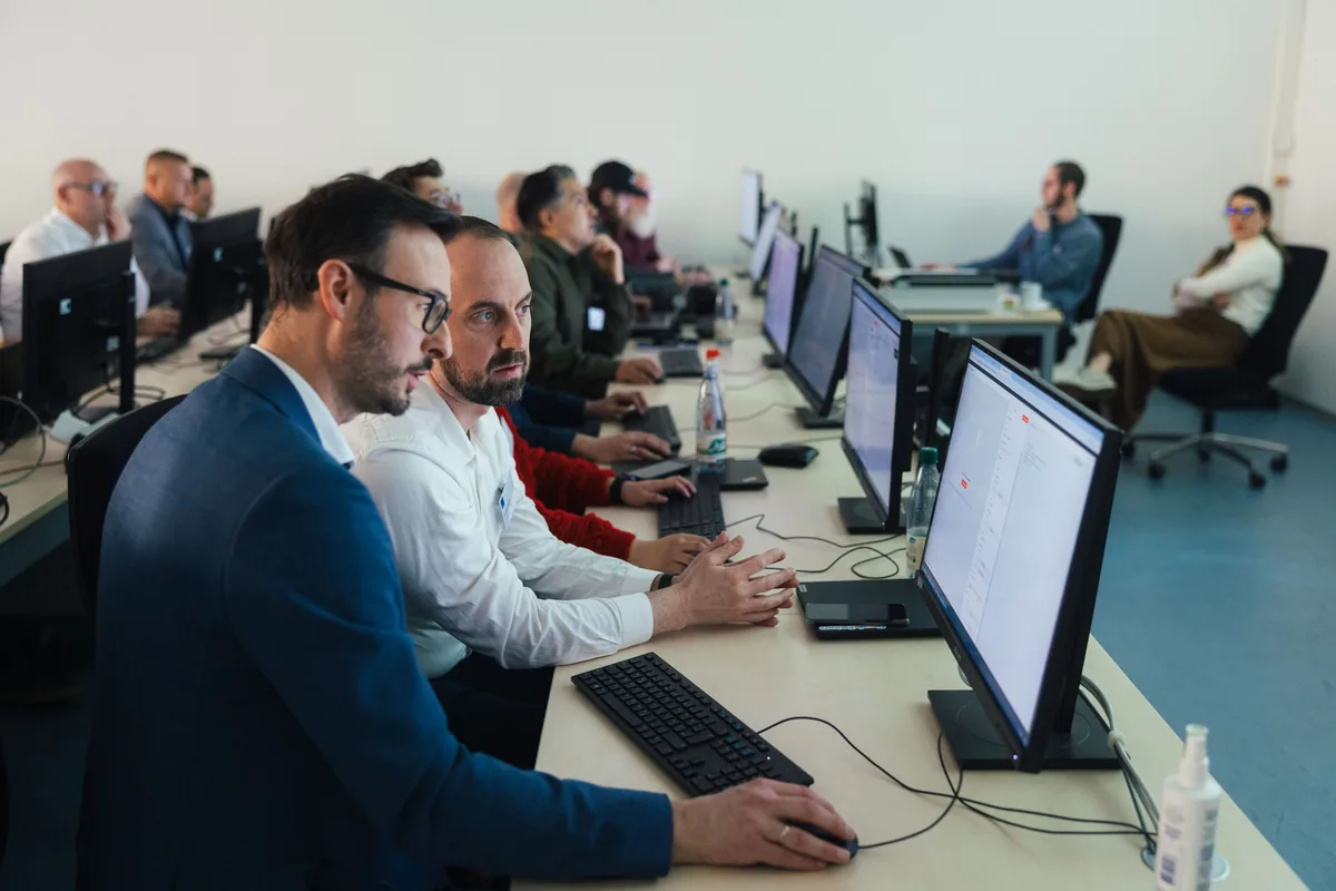 Zwei Männer in blauem Sakko und weißem Hemd schauen konzentriert auf einen Monitor, hinter ihnen die Reihen des Computerraums