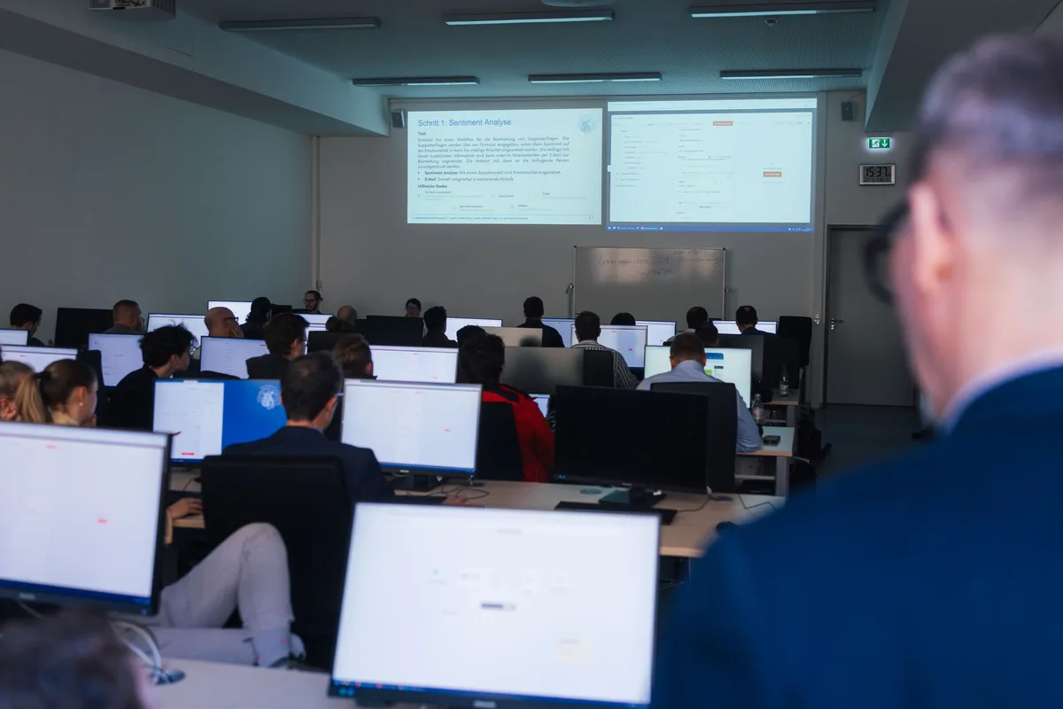 Blick über die Schulter eines Mannes auf den Raum voller Monitore, an der Wand zwei Projektionen mit Aufgabentext
