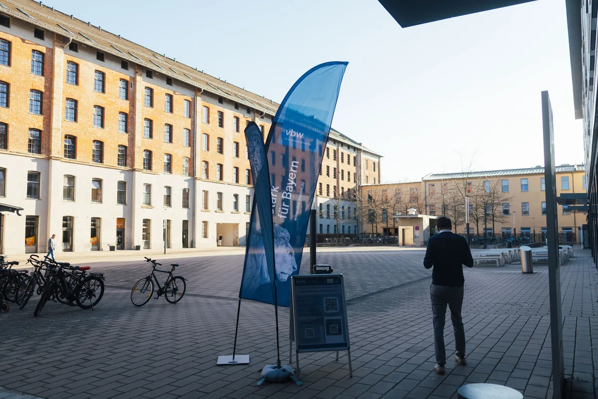 Platz mit hohem Backsteinbau im Hintergrund, im Vordergrund eine blaue Fahne, abgestellte Fahrräder und ein Mann von hinten