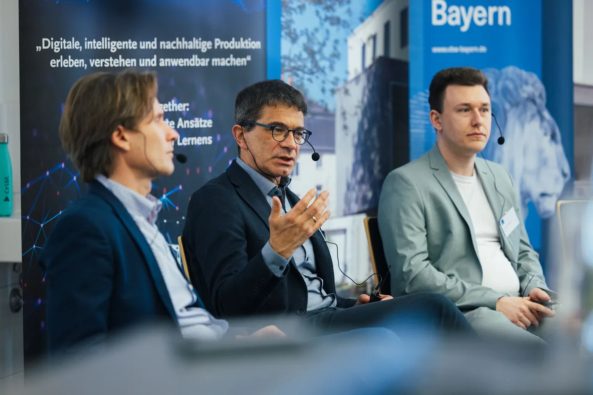 Mann mit Brille in der Mitte des Podiums gestikuliert mit offener Hand, rechts der Mann im grünen Sakko
