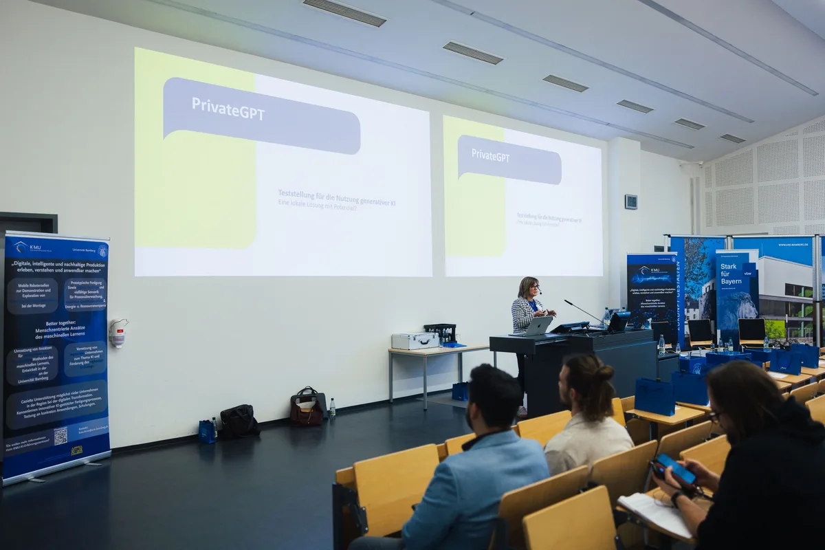 Frau am Rednerpult vor zwei Projektionen mit dem Titel „PrivateGPT“, im Vordergrund Zuhörer, einer schaut aufs Handy