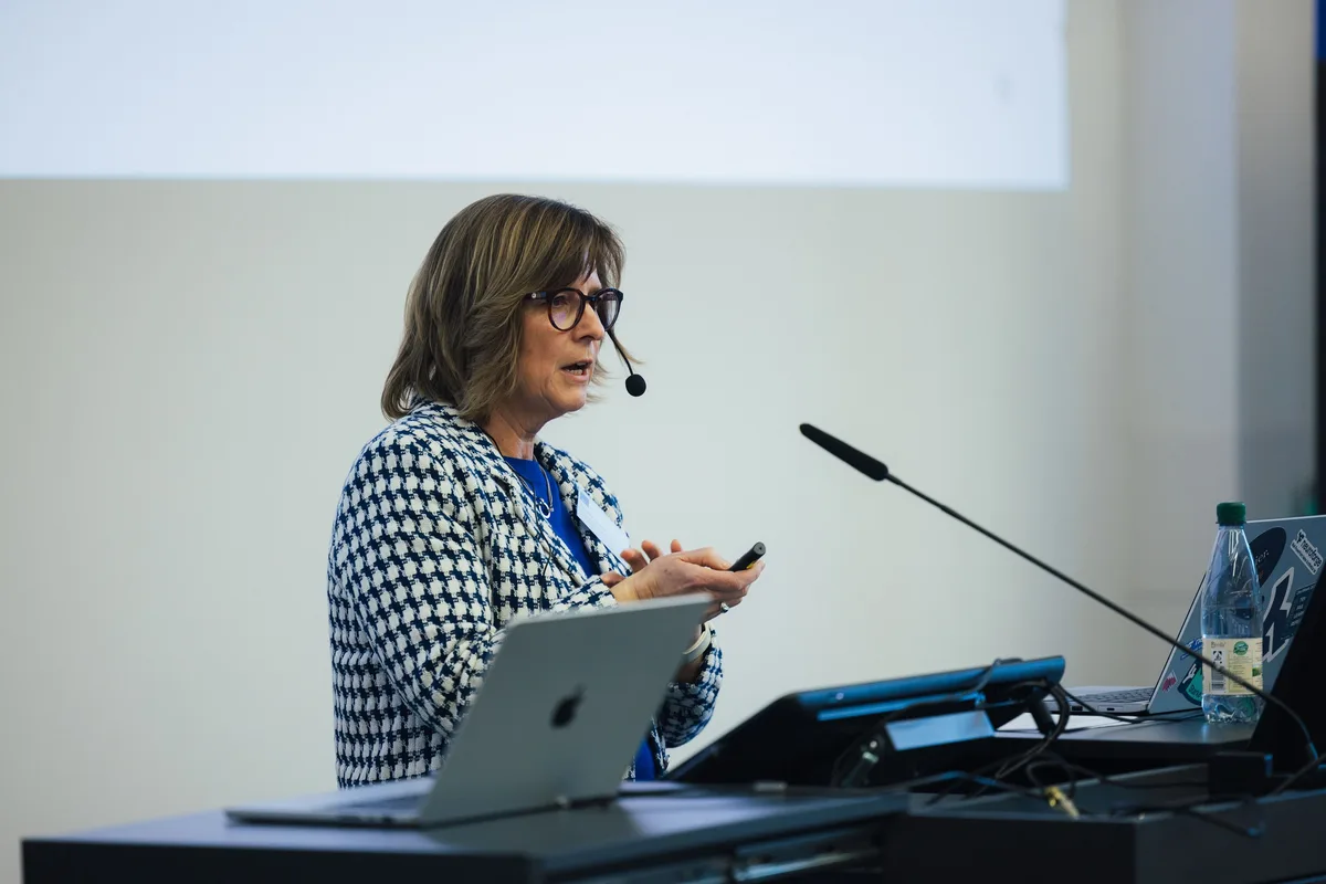 Frau im Hahnentrittmuster mit Headset spricht und hält einen Presenter, vor ihr ein aufgeklappter Laptop