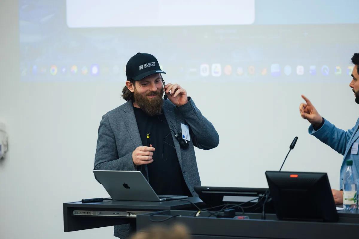 Mann mit Basecap und Bart im grauen Sakko lächelt am Pult, eine Hand am Headset, rechts zeigt ein Mann im Anschnitt nach oben