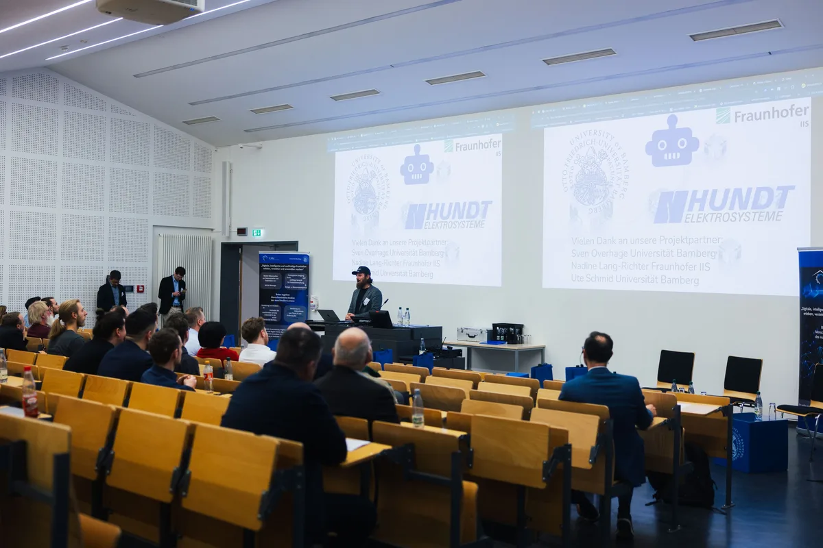 Blick über die Reihen im Hörsaal auf den Redner mit Cap am Pult, zwei Projektionen zeigen Logos von Projektpartnern